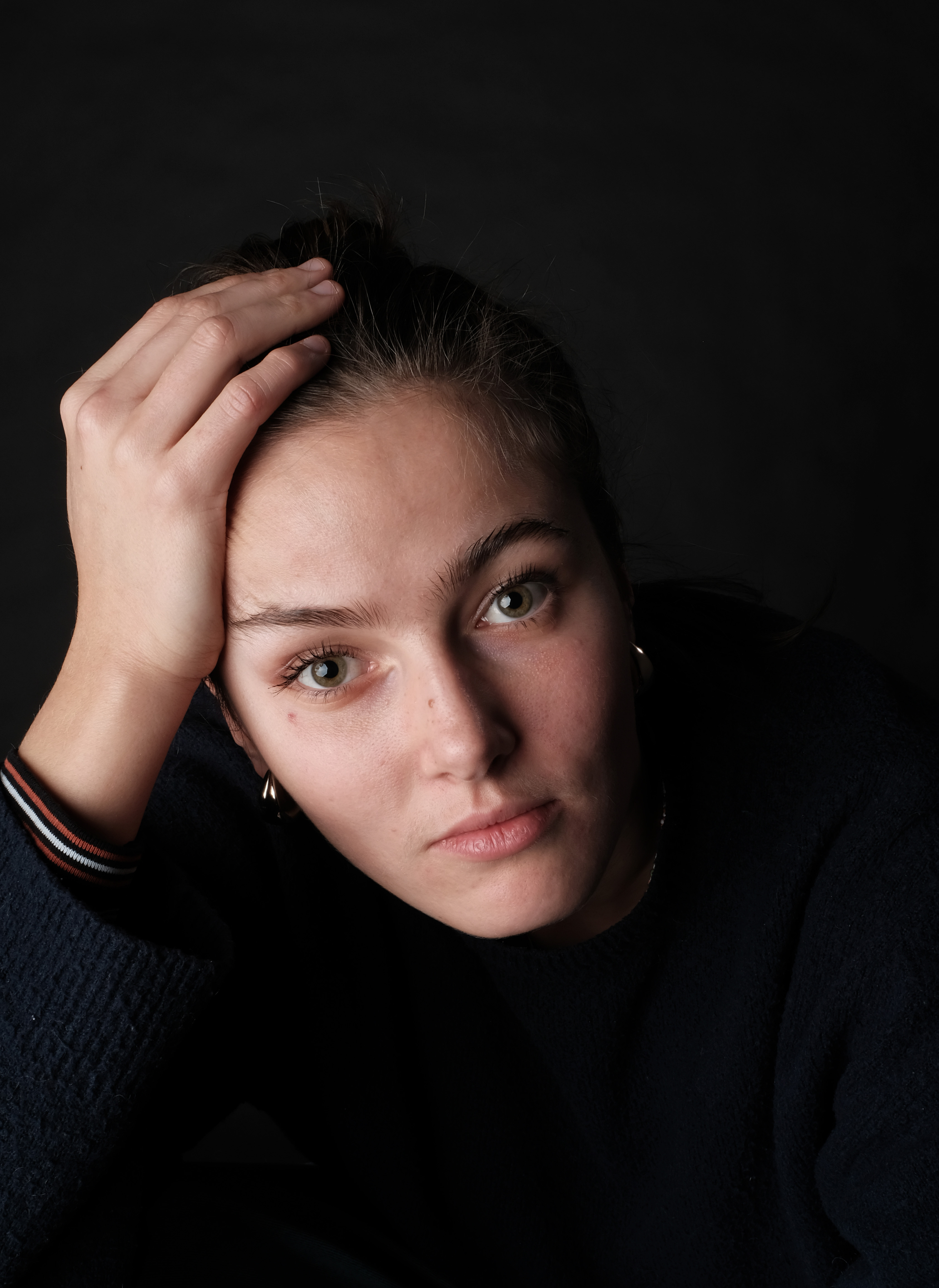 Portrait of a young woman with dark blond hair, in a bun. She's wearung a dark blue jumper and golden earrings