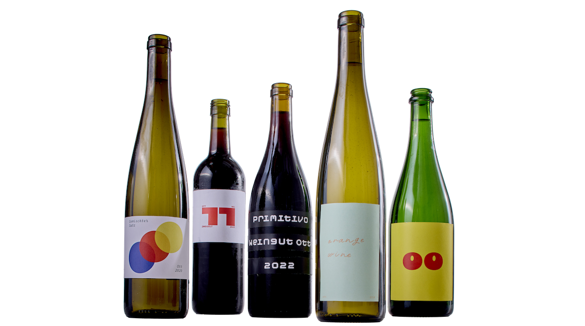 five wine bottles with colourful labels: the 1st bottle from the left is green and long, it has a white label with three circles arranged diagonally on it. the circles are blue, red and yellow and a little transparent. The font is small an put unto the upper left- and the lower right corner. The 2nd bottle from the left is dark red, almost black. It also has a white label and a red, bold 11 on it. The rest of the font is arranged around the 11. The 3rd bottle is also dark red, almost black and round-bodied, it has thee black banners on it, with a white, typewriter-like font. The 4th bottle, like the 1st one is green and long, the label is pastell green with an orange, cursive font on it, it is narrow and long. The 5th bottle is green and round-bodied and has a yellow label on it that has two Os on it. All bottles have wine in them and no cork.