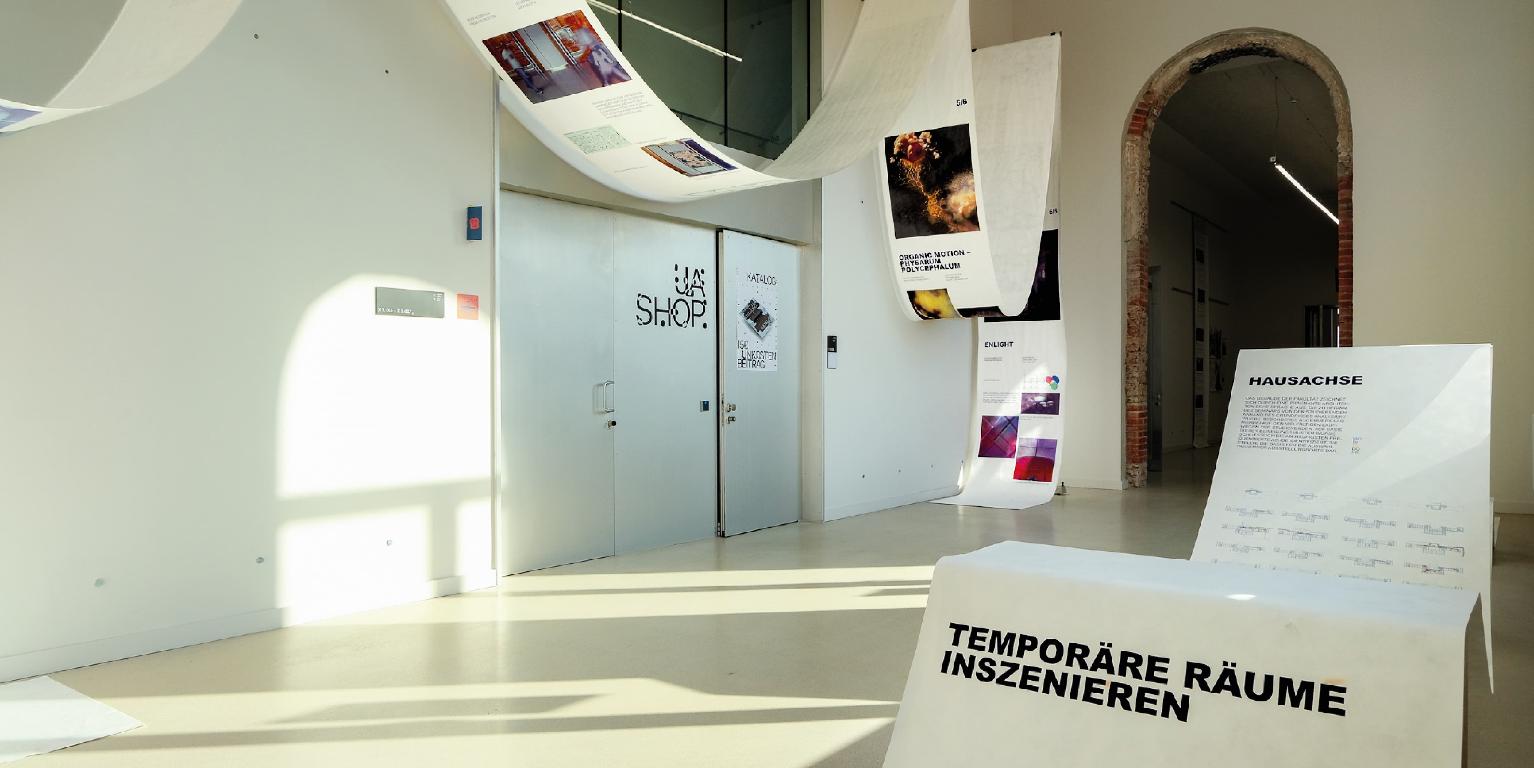 Two images, the lower one shows a wall, a big silver door and a silver ladder infront of that door. The upper image shows a hydraulic ramp with a young, dark blond man on it and a girl with dark hair that stands infront of it, with her back to the camera, shes wearing a white tshirt and a black, long skirt. A Banner is hanging above the hydraulic ramp